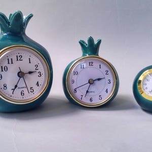 May include: Three green ceramic clocks shaped like pomegranates. The clocks have gold-colored trim and white faces with black hands. The largest clock is about 5 inches tall, the middle clock is about 4 inches tall, and the smallest clock is about 3 inches tall.