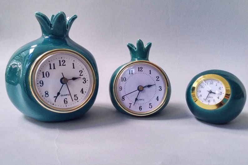 May include: Three green ceramic clocks shaped like pomegranates. The clocks have gold-colored trim and white faces with black hands. The largest clock is about 5 inches tall, the middle clock is about 4 inches tall, and the smallest clock is about 3 inches tall.