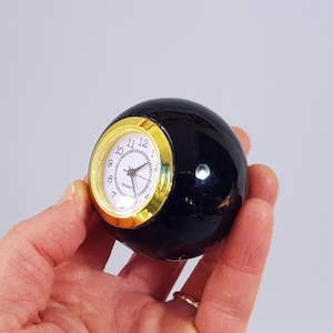 May include: A black sphere clock with a gold rim and white face. The clock face has black hands and black numbers.