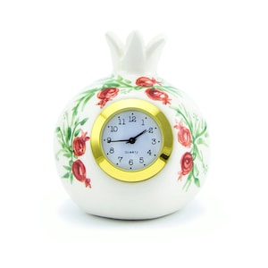 May include: White ceramic pomegranate clock with red and green painted details. The clock face has black hands and gold Roman numeral markings. The clock face reads "QUARTZ".
