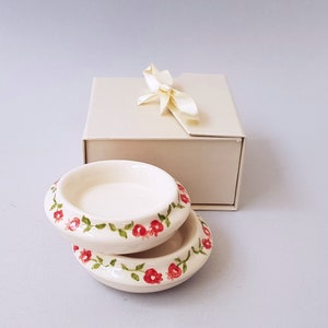 May include: Two small white ceramic dishes with a floral design. The dishes have a green vine pattern with red flowers. The dishes are stacked on top of each other and are in a white box with a cream-colored ribbon.