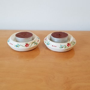 May include: Two white ceramic candle holders with floral designs in red, blue, and green. Each holder has a small, brown tea light candle inside.