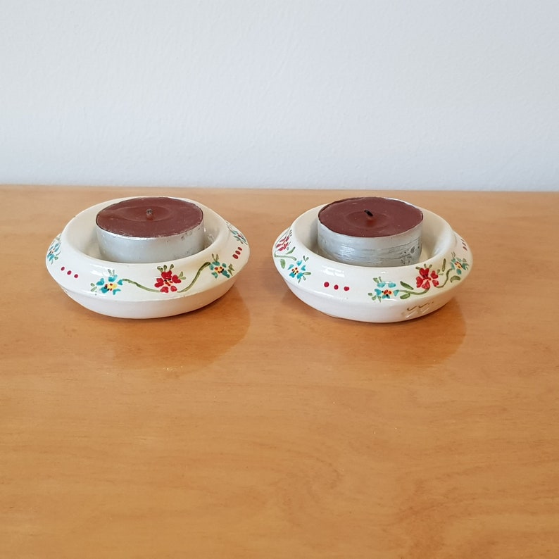 May include: Two white ceramic candle holders with floral designs in red, blue, and green. Each holder has a small, brown tea light candle inside.