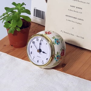 May include: White ceramic clock with gold trim and floral design. The clock face is white with black numbers and hands. The clock is set to 11:55.