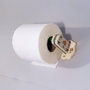 May include: White toilet paper roll on a ceramic toilet paper holder with a floral design. The holder is painted in shades of cream, green, and yellow.