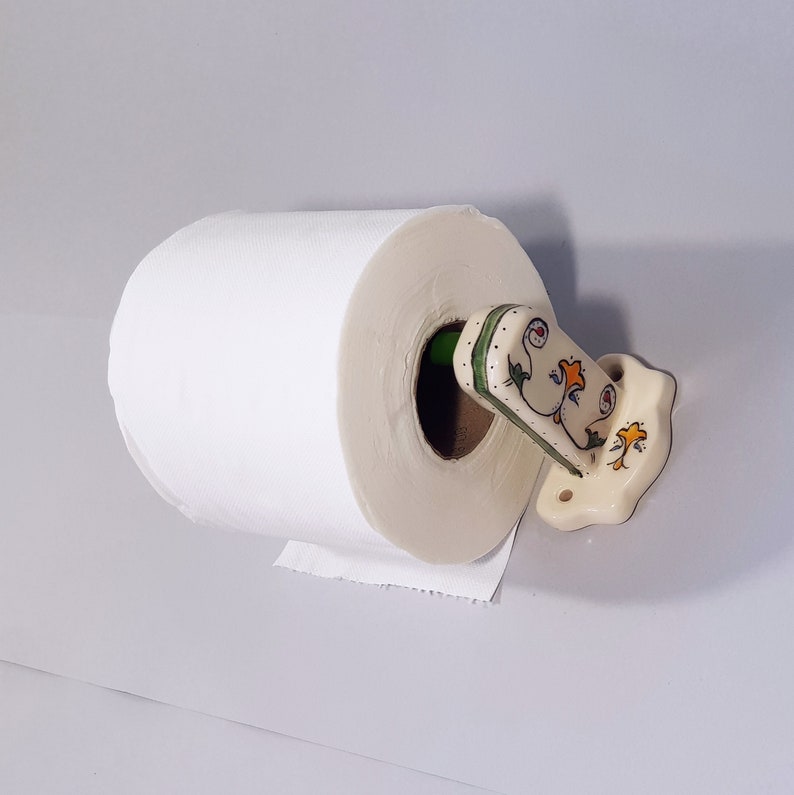 May include: White toilet paper roll on a ceramic toilet paper holder with a floral design. The holder is painted in shades of cream, green, and yellow.