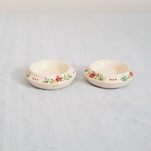 May include: Two small white ceramic bowls with a floral design. The bowls have a red, green, and blue floral pattern around the rim.