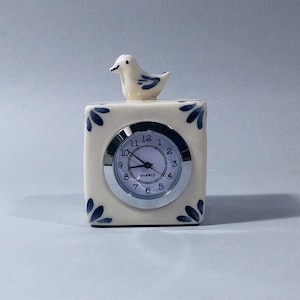 Puede incluir: Reloj de cerámica blanca con un diseño Delft azul y blanco. El reloj tiene un pequeño pájaro en la parte superior y una esfera redonda con agujas negras y números negros. La esfera del reloj dice "Quartz".