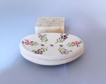 May include: White ceramic soap dish with a floral design in shades of pink, purple, and green. The dish has three drainage holes and is oval shaped.