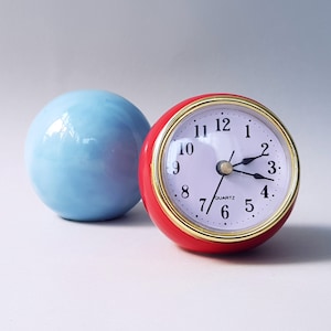 May include: A red clock with a white face and black hands. The clock is set to 10:10. The clock is sitting next to a light blue sphere.