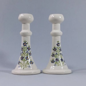 May include: A pair of white ceramic candle holders with a floral design. The flowers are blue, green, and yellow.