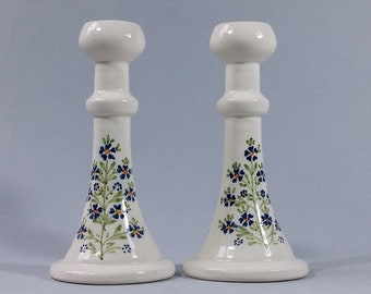 May include: A pair of white ceramic candle holders with a floral design. The flowers are blue, green, and yellow.