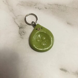 Peut inclure: Un porte-clés vert en forme de goutte avec une finition lisse et brillante. La surface présente trois protubérances circulaires en relief rappelant des petits pois et un visage simple avec deux yeux noirs et une petite bouche rose.