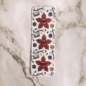 May include: White bookmark with a repeating pattern of Demogorgon flowers, cassette tapes, dice, and string lights. The bookmark is rectangular with rounded corners. The Demogorgon flowers are red, and the cassette tapes are blue and white.