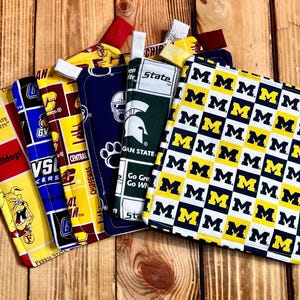 May include: A collection of fabric items featuring college team logos and colors. Includes a yellow and blue patterned item with the letter "M", and items with logos for Ferris State University, Central Michigan, and Michigan State.
