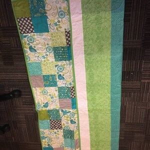 Pode incluir: Um edredom feito &agrave; m&atilde;o com um design de patchwork com quadrados de tecidos turquesa, verdes e castanhos. O edredom tem uma faixa branca no centro e uma borda verde lisa. O edredom tem aproximadamente 152 cm de comprimento.