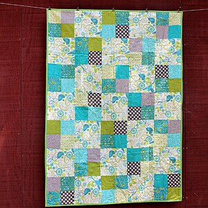 May include: A patchwork quilt with a green border, featuring squares of various patterns and colors. The quilt includes squares of teal, green, white with floral designs, and brown polka dots. The quilt is hanging against a red wooden background.