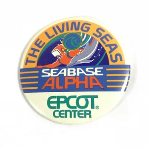 Puede incluir: Un botón redondo de color naranja, azul y blanco con el texto "The Living Seas Seabase Alpha Epcot Center" y un gráfico de un buzo y un delfín.