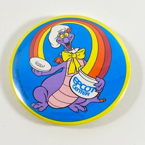 Könnte beinhalten: Ein lila Drache mit Kochmütze und gelber Schleife hält einen Teller und ein Glas, auf dem "EPCOT CENTER" steht. Der Drache steht vor einem Regenbogen auf blauem Hintergrund.