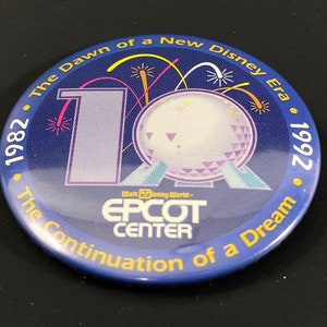 Op de afbeelding: Een blauwe knop met een gouden rand en tekst die luidt "1982 • The Dawn of a New Disney Era • 1992 • The Continuation of a Dream". De knop heeft een witte en paarse Spaceship Earth met vuurwerk op de achtergrond. De tekst "Walt Disney World EPCOT CENTER" staat ook op de knop.