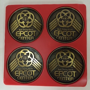 May include: Four gold-colored metal coasters with the text "Walt Disney World EPCOT CENTER" on each. The coasters are arranged in a square pattern on a red surface.