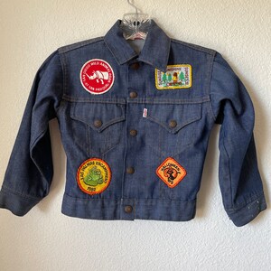 May include: A vintage blue denim jacket with multiple patches. Patches include a rhino, a frog, and a Halloween design. The jacket has button closures and two chest pockets. The Levi's brand tag is visible.