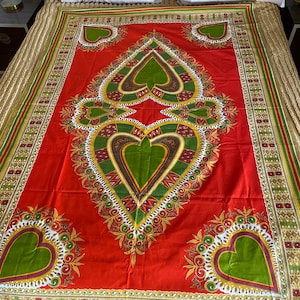 May include: A red textile with a large green heart-shaped design in the center and smaller heart shapes in the corners. The design includes gold, white, and brown accents. The textile is likely a bedspread or tablecloth.