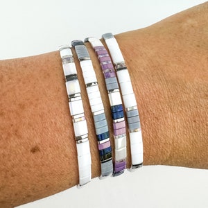 May include: Four stackable bracelets with white, gray, and purple tiles. The bracelets are made of ceramic and have a matte finish.