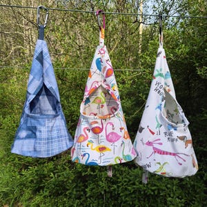 May include: Three hanging fabric pet hammocks. One is blue plaid, another features flamingos, and the third has a dog-themed print with the words "Woof Woof" and "Ruff Ruff". Each hammock is suspended by a carabiner.
