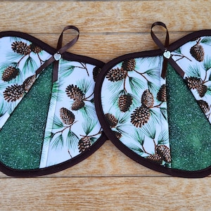 May include: Two heart-shaped pot holders with a brown trim and hanging loop. The fabric features a pine cone and pine needle design on a white background. A green triangle is sewn into the design. The pot holders are on a wooden surface.