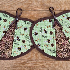 May include: Two heart-shaped pot holders with a brown trim and hanging loop. The pot holders feature a light green gingham pattern with bees and pinecones, and a brown floral pattern. The pot holders are on a wooden surface.