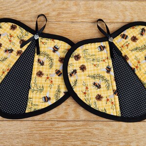 May include: Two heart-shaped pot holders with a bee and pinecone pattern on a yellow checkered background. One section is black with white polka dots. Each has a black trim and a small black ribbon.