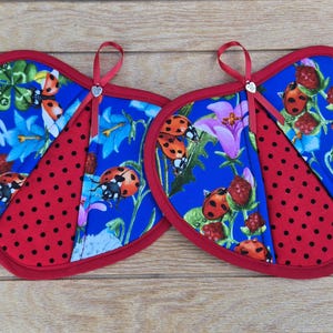 May include: Two heart-shaped potholders. The potholders have a blue floral pattern with ladybugs and a red polka dot accent. They are trimmed in red and have a red ribbon loop for hanging.