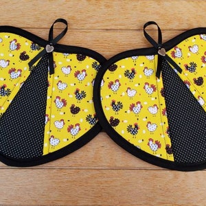 May include: Two heart-shaped potholders. Each potholder has a yellow background with a chicken pattern, a black polka dot section, and a black border. A black ribbon and a small heart charm are attached to each potholder.