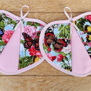May include: Two heart-shaped pink pot holders with a floral pattern featuring butterflies. The pot holders have a light blue background with pink roses and green leaves. The butterflies are in various colors, including blue, yellow, orange, and brown.