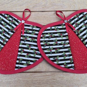 May include: Two heart-shaped pot holders featuring a black and white striped pattern with a holly leaf and berry print. Each piece has a solid red glittery fabric panel, a red fabric border, and a small red ribbon loop with a silver-toned charm.
