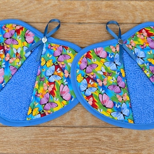 May include: Two heart-shaped pot holders with a vibrant butterfly print in shades of pink, yellow, and blue. Each pot holder has a blue border and a blue fabric section. A blue ribbon loop is attached for hanging.