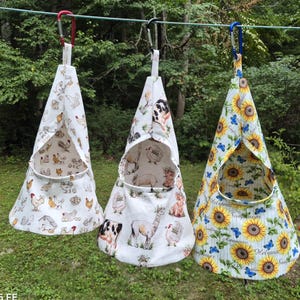 May include: Three cone-shaped fabric hanging storage bags suspended from a clothesline by red, black, and blue carabiners. The bags feature prints of chickens, farm animals including a cow and alpaca, and sunflowers with blue butterflies respectively.