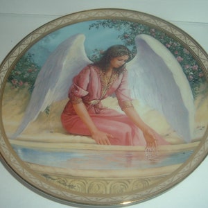May include: A porcelain plate with a gold rim features a painting of an angel with white wings sitting by a pool of water. The angel is wearing a pink robe and has long brown hair. The background is a garden with green trees and pink flowers.