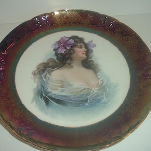 May include: A vintage porcelain plate with a raised, iridescent, burgundy and gold rim. The centre of the plate features a painted image of a woman with long brown hair wearing a blue and white dress and a flower crown.