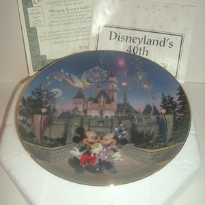 May include: A decorative plate featuring Mickey and Minnie Mouse in front of Sleeping Beauty's Castle at Disneyland. The plate has a gold rim and includes fireworks and Tinkerbell. The plate is part of the Disneyland 40th Anniversary collection.