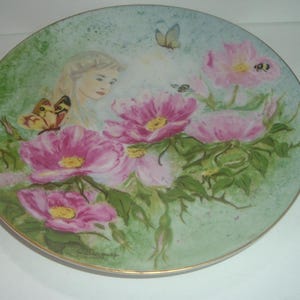 Puede incluir: Un plato decorativo con un diseño pintado de una niña con cabello rubio y una mariposa, rodeada de flores rosas y hojas verdes.