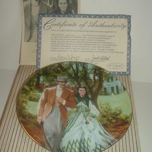 Pode incluir: Um prato de porcelana colecionável que apresenta uma cena do filme E o Vento Levou. O prato retrata Scarlett O'Hara e Rhett Butler caminhando juntos em uma paisagem verde. O prato é emoldurado com uma borda dourada e tem um certificado de autenticidade.