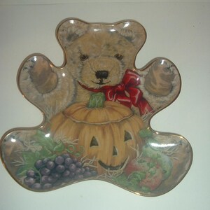 Puede incluir: Un plato decorativo con forma de oso con un borde dorado. El plato presenta un oso de peluche, una calabaza de Halloween, uvas y manzanas. El oso tiene un lazo rojo. El plato es un artículo decorativo con temática de otoño o Halloween.
