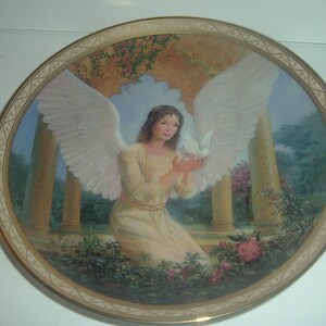 Può includere: Un piatto in porcellana con un bordo dorato presenta un dipinto di un angelo con ali bianche che tiene una colomba bianca. L'angelo indossa un abito bianco e ha lunghi capelli castani. Lo sfondo è un giardino con alberi verdi e fiori.