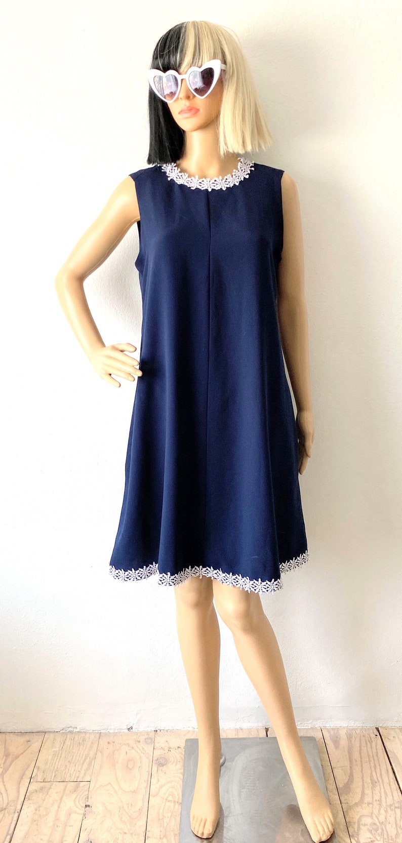 May include: A navy blue sleeveless dress with white lace trim around the neckline and hem. The dress is knee-length and has a simple, A-line silhouette.