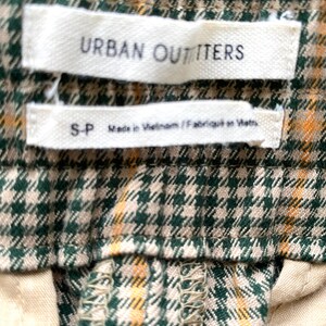 May include: Close-up of a green and tan plaid fabric with two white labels. The top label reads "URBAN OUTFITTERS". The bottom label reads "S-P Made in Vietnam / Fabriqu&eacute; en Vietnam."