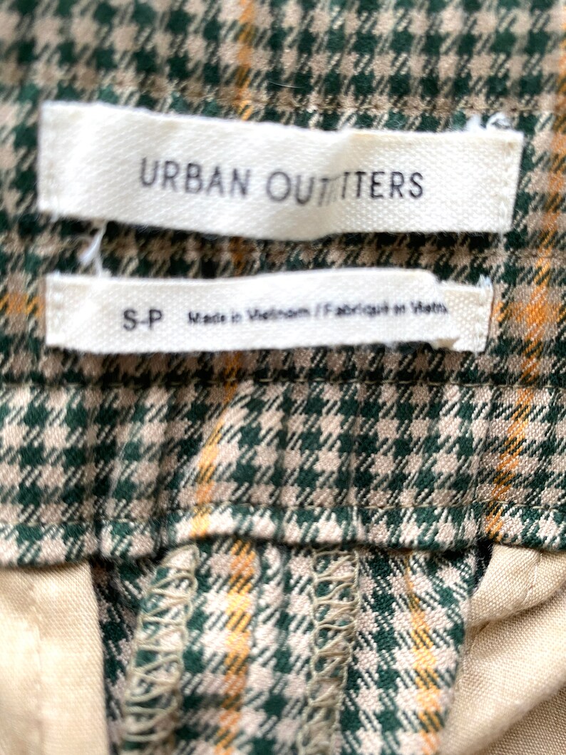 May include: Close-up of a green and tan plaid fabric with two white labels. The top label reads "URBAN OUTFITTERS". The bottom label reads "S-P Made in Vietnam / Fabriqu&eacute; en Vietnam."