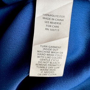 May include: A white care label with black text on a blue fabric background. The label provides instructions for washing and drying a garment made of 100% polyester. The label states "Turn garment inside out, machine wash or hand wash cold with like colors, gentle cycle, do not bleach, remove promptly, tumble dry low, warm iron if needed, or dry clean". The label also includes the RN number 103715.