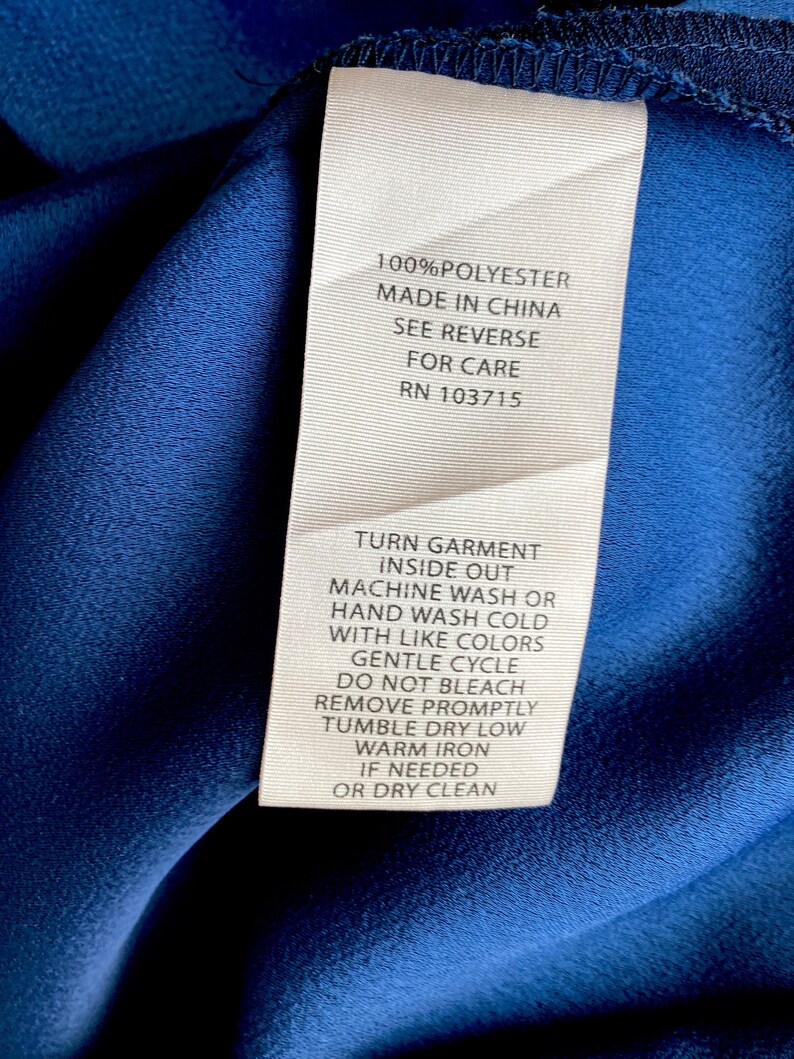 May include: A white care label with black text on a blue fabric background. The label provides instructions for washing and drying a garment made of 100% polyester. The label states "Turn garment inside out, machine wash or hand wash cold with like colors, gentle cycle, do not bleach, remove promptly, tumble dry low, warm iron if needed, or dry clean". The label also includes the RN number 103715.
